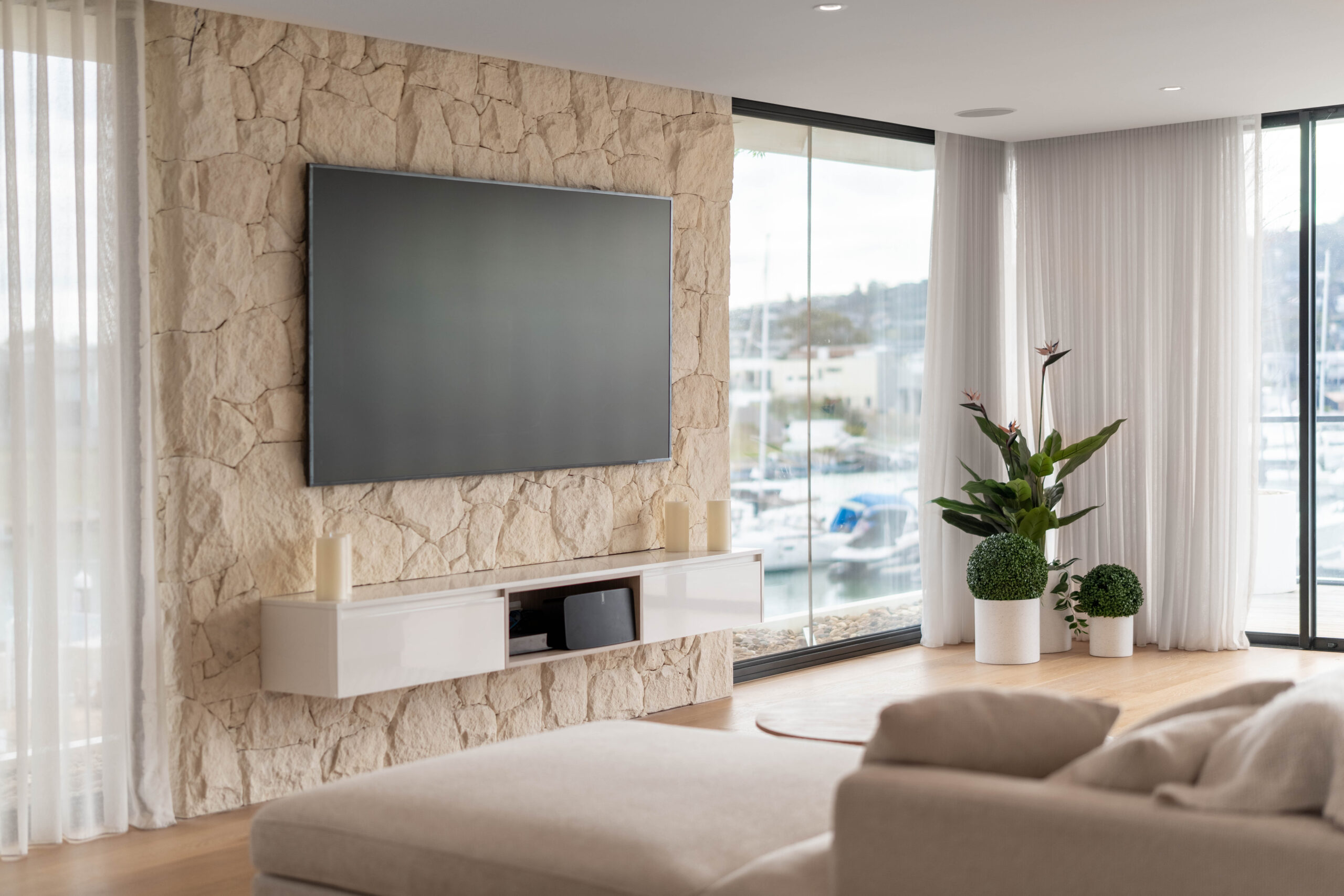 Living room with stone feature wall, wall-mounted television, floating cabinetry and full-height glass doors overlooking a marina