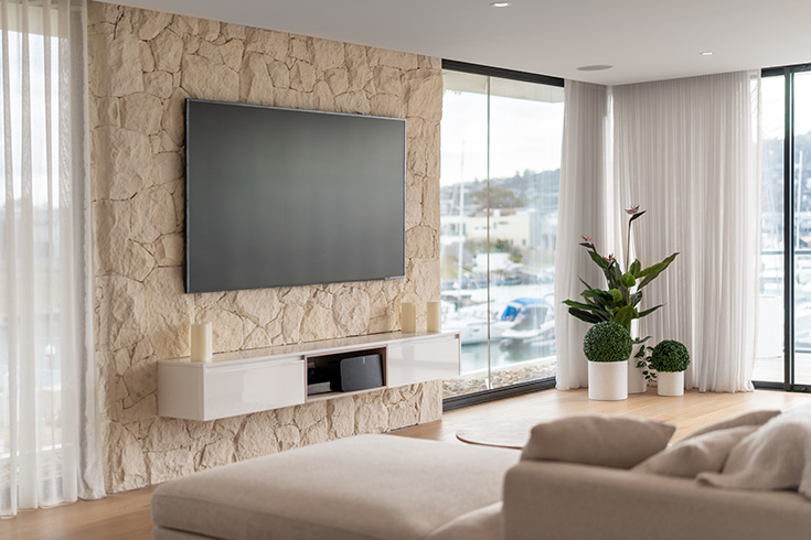 Luxurious stone feature wall, wall-mounted television, sleek custom entertainment cabinets, and black steel corner floor-to-ceiling windows.